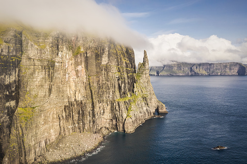 Faroe Islands Trollkonufingur Sea Stack Rock - Monolith - also called 'The Troll Woman's Finger' or 'Witch Finger' on the island of Vagar under moody low hanging cloudscape in summer. Aerial Drone Point of View of Vágar Island South-East Coast with Trøllkonufingur Sea Stack the famous 313m tall Monolith at the Vagar fjord coastline. Eysturoy Island coast in the background. East Coast Fjord of Sandavagur village, Vágar Island, Faroe Islands, Kingdom of Denmark, Nordic Countries, Scandinavia, Europe