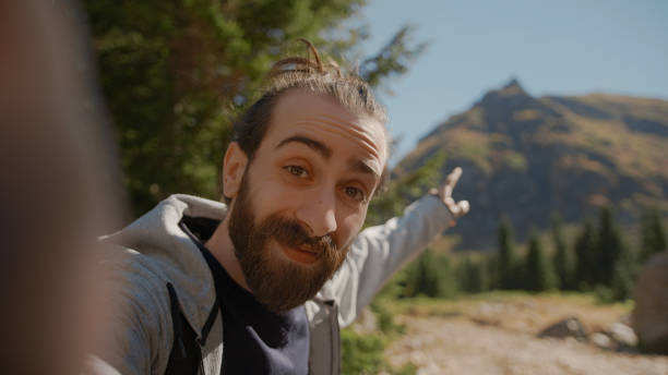 Hippie climber with his backpack behind him, with his handheld camera or phone, salutes the audience or the person he is talking and points to a high mountain. vlogger or video chat in nature concept