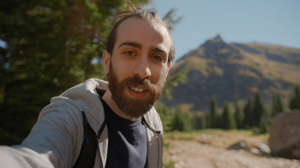 istock Hippie climber with his backpack behind him, with his handheld camera or phone, salutes the audience or the person he is talking, vlogger or video chat in nature concept 1280738548