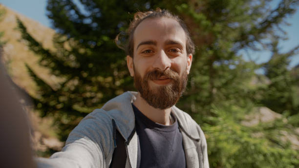 Smile face hippie climber with his backpack behind him, with his handheld camera or phone, salutes the audience or the person he is talking, vlogger or video chat in nature concept