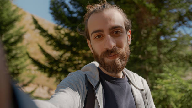 istock Hippie climber with his backpack behind him, with his handheld camera or phone, salutes the audience or the person he is talking, vlogger or video chat in nature concept 1280740126