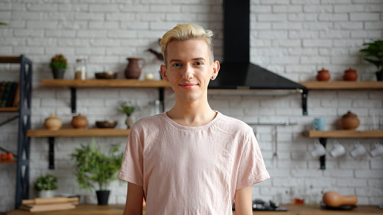 Attractive young guy with blond colored hair wearing pink casual t-shirt smiles to camera standing in contemporary kitchen