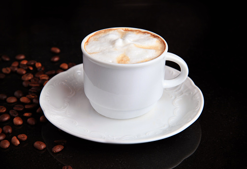 A cup of cappuccino on a white plate and a table with coffee beans