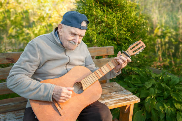 87 year-old mustachioed man plays an acoustic guitar outdoors in the garden. old man of caucasian nationality remembers his youth and uses a musical instrument in nature - 80 year old imagens e fotografias de stock