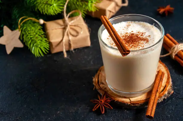 Christmas Hot Drink. Eggnog with Cinnamon in Glass with Branches Fir Tree on Dark Background.