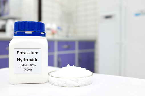 Selective focus of a bottle of pure potassium hydroxide or KOH chemical compound beside a petri dish with white solid pellets. Chemistry research laboratory background with copy space.