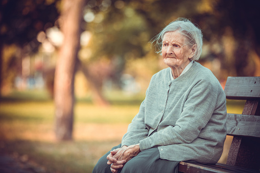 Portrait of senior woman sitting on bench in autumn park. Old lady feeling lonely and sad. Frustrated aged female outdoors.