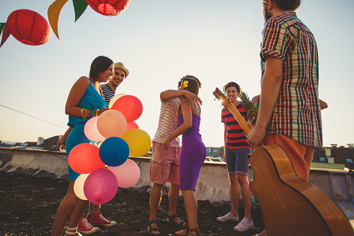 Wide shot of group of friends in colorful outfits hugging each other while bringing balloons and a guitar for a rooftop party during sunny days.