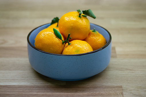 Blue bowl with mandarin oranges with green leaf. Ripe sour citrus fruit on a wooden table close-up