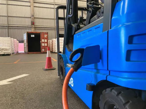 Photo of Electrical forklift vehicle park and plug in for charging battery inside of logistic warehouse. Alternative energy source for car and machine in industrial business segment. Automotive technology.