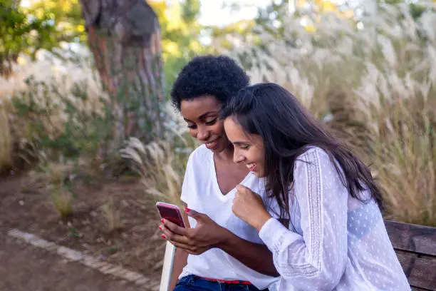 Two female friends sitting on a bench interacting with a mobile phone