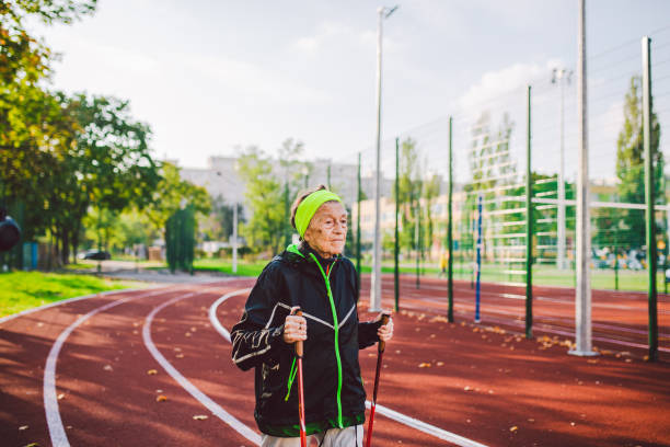 Old woman in sportswear practicing nordic walking outdoors on rubber treadmill in stadium. Older female walk by scandinavian walk use trekking sticks and nordic poles. Retired people healthy lifestyle. anciana en ropa deportiva practicando marcha nórdica al aire libre en cinta de correr de goma en el estadio. el paseo femenino más antiguo por caminata escandinava utiliza bastones de trekking y bastones nórdicos. personas jubiladas estilo de vida salud