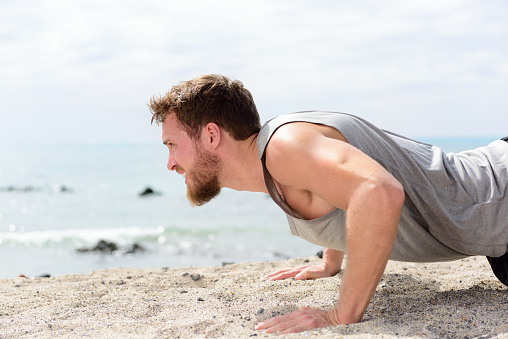 Fitness man doing push-up exercise on beach. Portrait of fit guy working out his arm muscles and body core with pushup exercises on sand beach.
