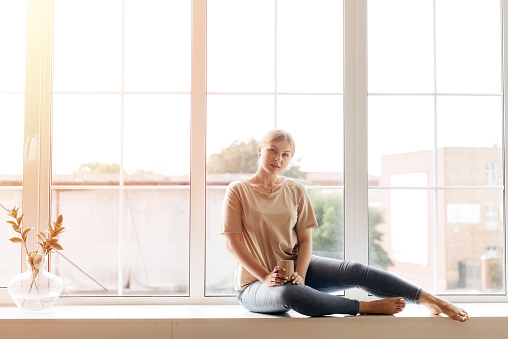 Beautiful view. Attractive young woman sitting on the windowsill and looking to the camera