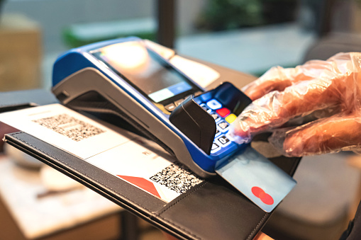 Cropped shot of an Asian male hands with protective gloves pressing button from the portable payment machine with inserted debit card during Covid 19 pandemic in the restaurant