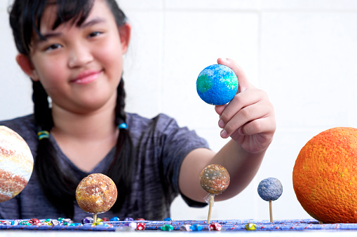 Focus at girl hand holding earth planet with blurred background, young smiling Asian girl showing her homemade solar system model at home