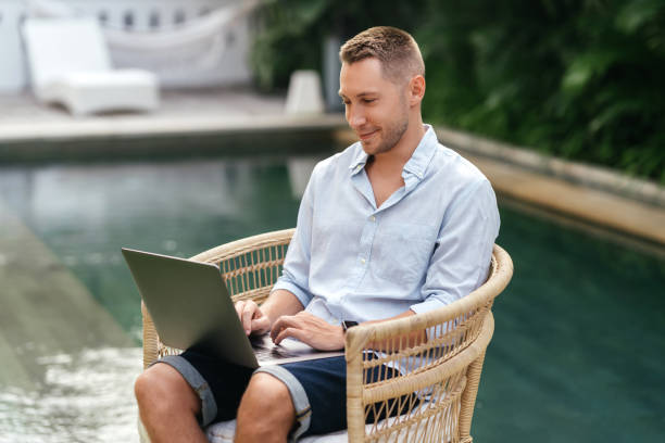 istock Young man using a laptop computer in a garden with a swimming pool. 1280840347