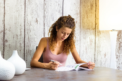 Attractive young woman working on crossword puzzles at home in a magazine seated at a table with copyspace