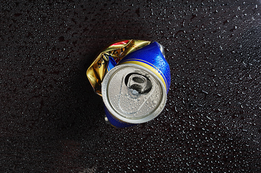 Crumpled beer can in drops of water on black background, top view