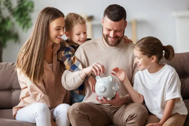 Happy family: cheerful mother and father with kids smiling and putting coins into piggy bank while sitting on sofa at home