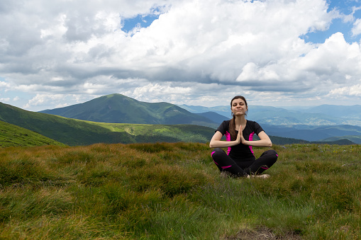 A beautiful girl sits in a meadow with her hands folded in front of her, among the beautiful mountains
