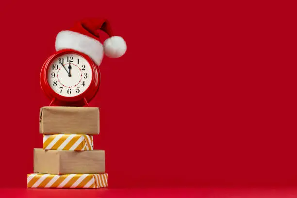 Red alarm clock in Santa Claus hat on gift boxes. Red background. Concept of coming Christmas and New Year, holiday sales. Space for text