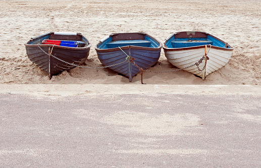 A trio of clinker built wooden rowing boats rest, chained together on the sandy beach, at the waters edge alongside the waterfront promenade Bournemouth, England