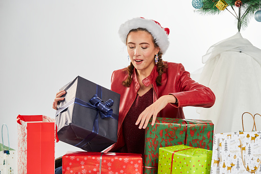 Young brunette Spanish woman dressed in Santa hat celebrating Christmas with gifts and decorating the Christmas tree, during photo shoot indoors with white background