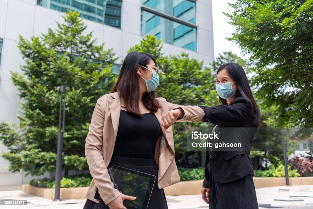 Business colleagues wearing face masks greeting with elbows during coronavirus pandemic Two Asian businesswomen greeting by bumping elbows outside of office, new normal handshake during covid-19 pandemic. Singapore Stock Photo