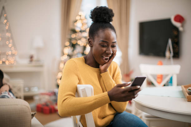 Staying in touch with friends and family during winter holidays Happy young African American woman using a smart phone, staying in touch with her family and friends and wishing them a Marry Christmas and a happy new year christmas excitement stock pictures, royalty-free photos & images