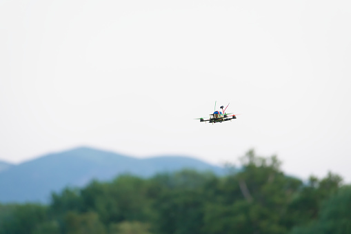 Colorful propellers lift this racing FPV quad copter drone through the air. Plain white sky background with blurred hills and trees beneath