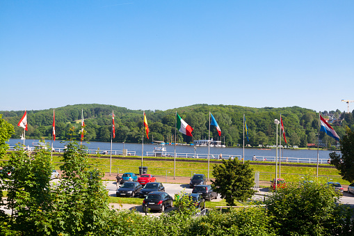 Parking lot and international flags at Lake Baldeneysee in Essen. Sorng season shot of lake and landscape. Cars are parked below lake. At opposite side are woods at Ruhr. Some people are in scene