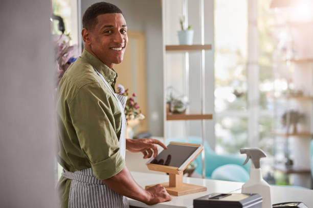 Smiling male shop assistant working at florist Waist up portrait of cheerful young handsome man selling flowers and standing at counter with tablet and pos terminal point of sale tablet stock pictures, royalty-free photos & images