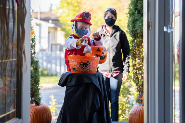 Young Boy wearing protective mask taking candies on Halloween at front door of a house Young Boy wearing protective mask taking candies in a self service candy basket on Halloween at front door of a house during COVID-19 pandemic. People are putting a candy basket outside to keep social distancing and let children celebrate Halloween and trick or treat. The little redhead boy is accompanied by his mom. candy house stock pictures, royalty-free photos & images