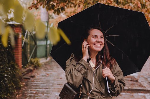 young pretty woman portrait listening music with headset while walking by street with umbrella under rain. copy space