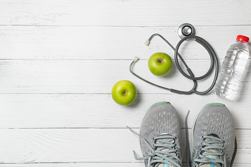 Running shoes with green apples and stethoscope near  fresh water bottle on  white wood background, exercise  and diet concept