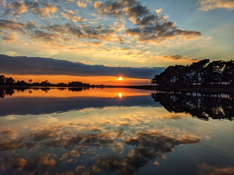 The sun falls towards the horizon over still water that reflects the banks of colours and clouds.