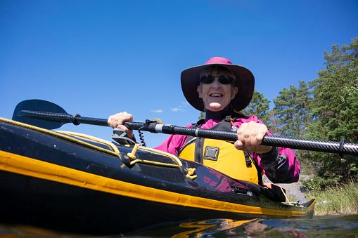 A mature woman sitting in a sea kayak, looking down at the camera.
The camera is in the water
She is wearing a purple paddle jacket and yellow PFD.