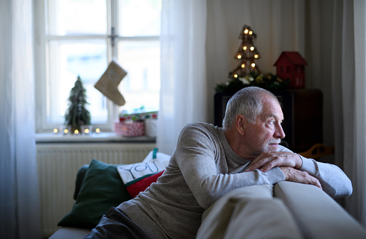Portrait of lonely senior man sitting on sofa indoors at Christmas, solitude concept.