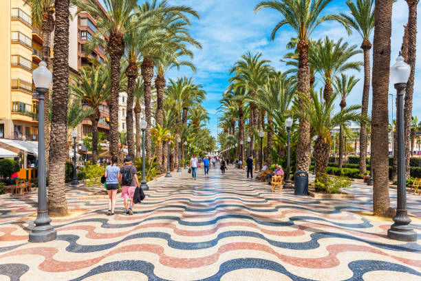 Alicante, Spain - March 29, 2018: People walking on a palm-lined promenade in Alicante, Spain. Alicante is a city located in the southeast of the Iberian Peninsula, on the shores of the Mediterranean Sea. It is the capital of the Alicante province and has about 330,000 inhabitants.