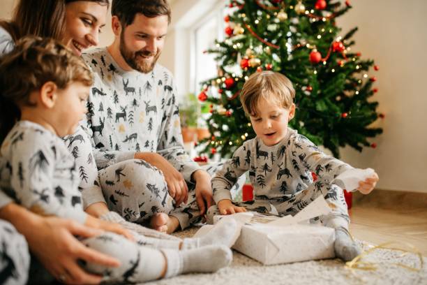 Family opening christmas presents Young family of four gathered together on a christmas morning wearing pajamas unwrapping christmas  gifts. They are smiling and having the best time. Warm colors, horizontal photo format. Bright and cozy living room with christmas tree in the background. Copy space. pajamas stock pictures, royalty-free photos & images