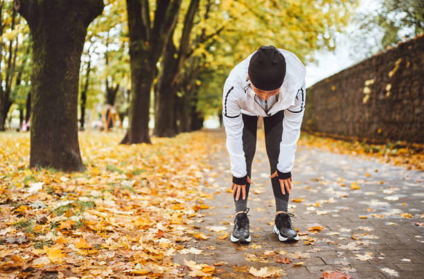 femme athlétique d’âge moyen a habillé des vêtements de course modernes restant et se penchent pour étirer le dos sur la passerelle après le jogging dans le parc de ville d’automne. active running people concept image. - se courber en avant photos et images de collection