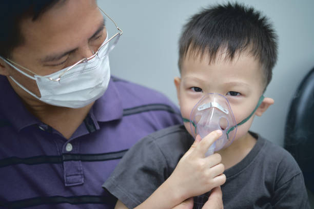 Asian father helping his toddler son with inhalation therapy by the mask of inhaler.  Little kid with respiratory problem with oxygen mask breathes through nebulizer Asian father helping his toddler son with inhalation therapy by the mask of inhaler. Sick smiling little kid with respiratory problem with oxygen mask breathes through nebulizer - Selective focus pediatric nebulizer mask stock pictures, royalty-free photos & images
