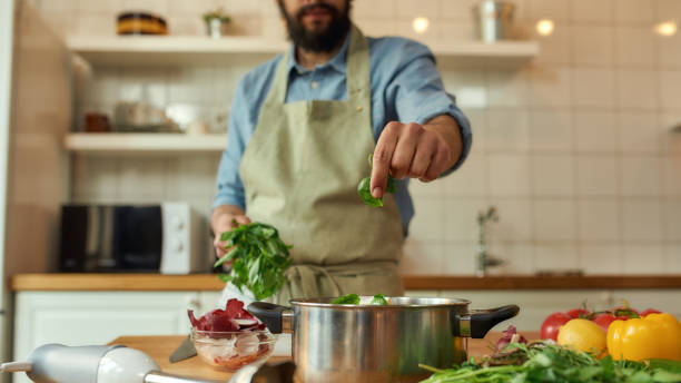 Cropped shot of man, chef cook adding basil leaf to the pot with chopped vegetables while preparing healthy meal, soup in the kitchen. Cooking at home, Italian cuisine concept