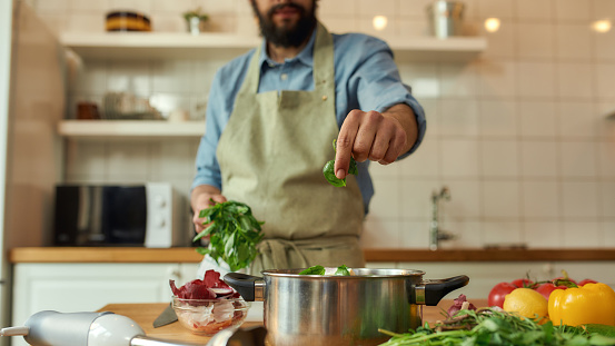 Cropped shot of man, chef cook adding basil leaf to the pot with chopped vegetables while preparing healthy meal, soup in the kitchen. Cooking at home, Italian cuisine. Focus on hand. Web Banner