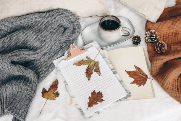 Fall breakfast still life. Cup of coffee, open book and notepad with colorful maple and oak leaves. White linen background. Linen pillows and grey sweater in bed, autumn lifestyle concept. Top view. el desayuno de otoño todavía es vida. taza de café, libro abierto y bloc de notas con coloridas hojas de arce y roble. fondo de lino blanco. almohadas de lino y suéter gris en la cama. concepto de estilo de vida de otoño. vista superior.