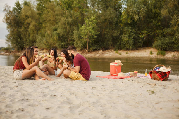 istock Group of five young people eating corn by the river at the beach picnic 1280973408