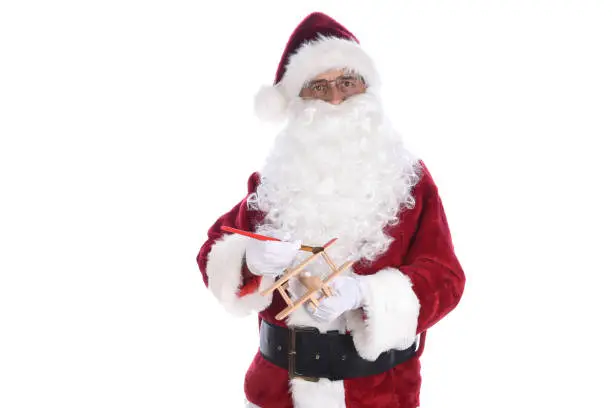 Senior man wearing a traditional Santa Claus costume painting a toy wooden airplane. Isolated on white.