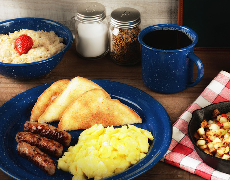 A country style scrambled egg breakfast on a rustic wooden restaurant table with oatmeal, potatoes and coffee with warm side light.