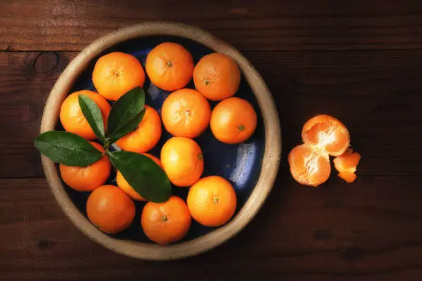 Flat lay image of a plate of fresh picked Mandarin Oranges in a rustic wood table with one peeled piece of fruit with warm side light.